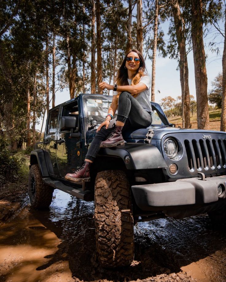 a woman is sitting on top of a jeep in the mud with trees behind her