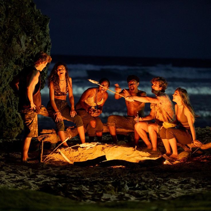 a group of people sitting around a campfire on the beach at night with torches