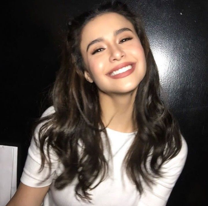 a woman with long brown hair and white shirt smiles at the camera while standing in front of a black wall