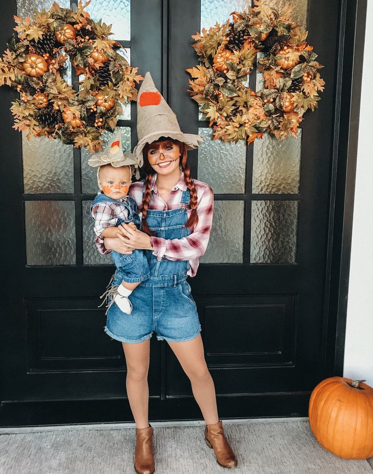 a woman holding a baby in front of a black door wearing a scarecrow hat