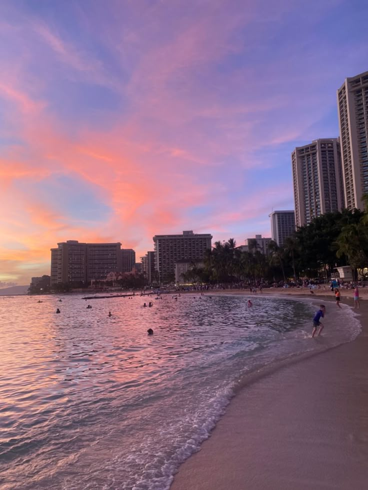 people are swimming in the ocean at sunset on a beach with high rise buildings behind them