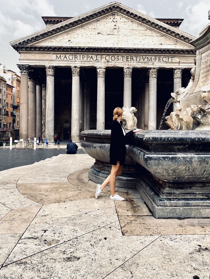 a woman standing in front of a building with columns and a fountain on the ground