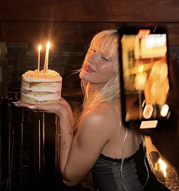 a woman holding a cake with two candles in front of her face while taking a selfie