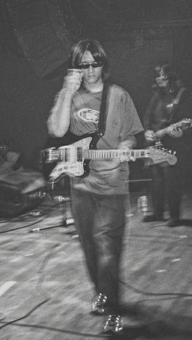 black and white photograph of man playing guitar on stage with other musicians in the background