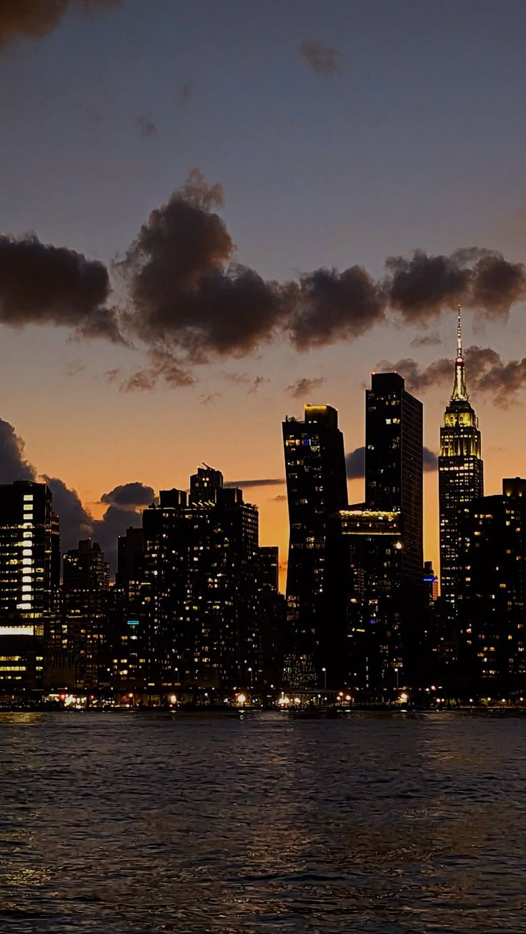 the city skyline is lit up at night as seen from across the water with clouds in the sky