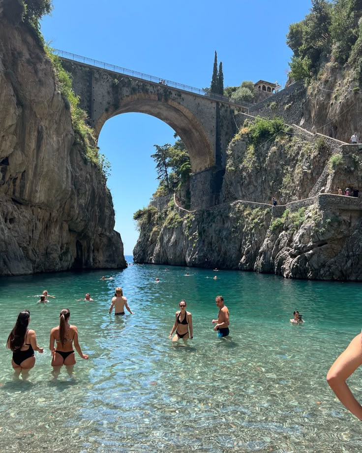 many people are swimming in the water near a stone bridge and some cliffs on either side