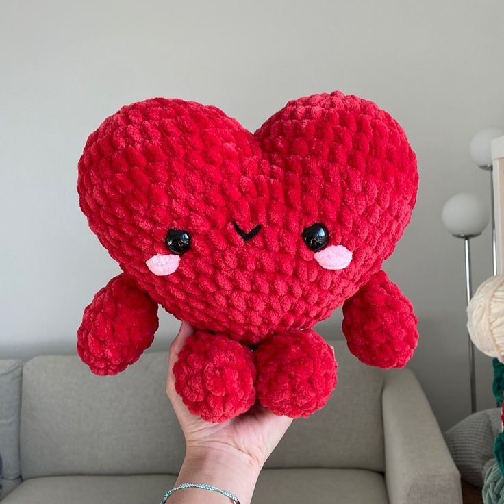 a person holding up a red heart shaped stuffed animal in front of a gray couch