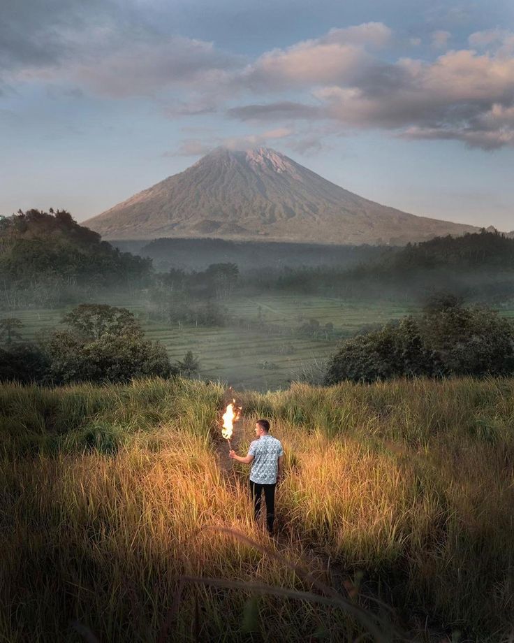 a man standing on top of a lush green field next to a tall mountain covered in clouds