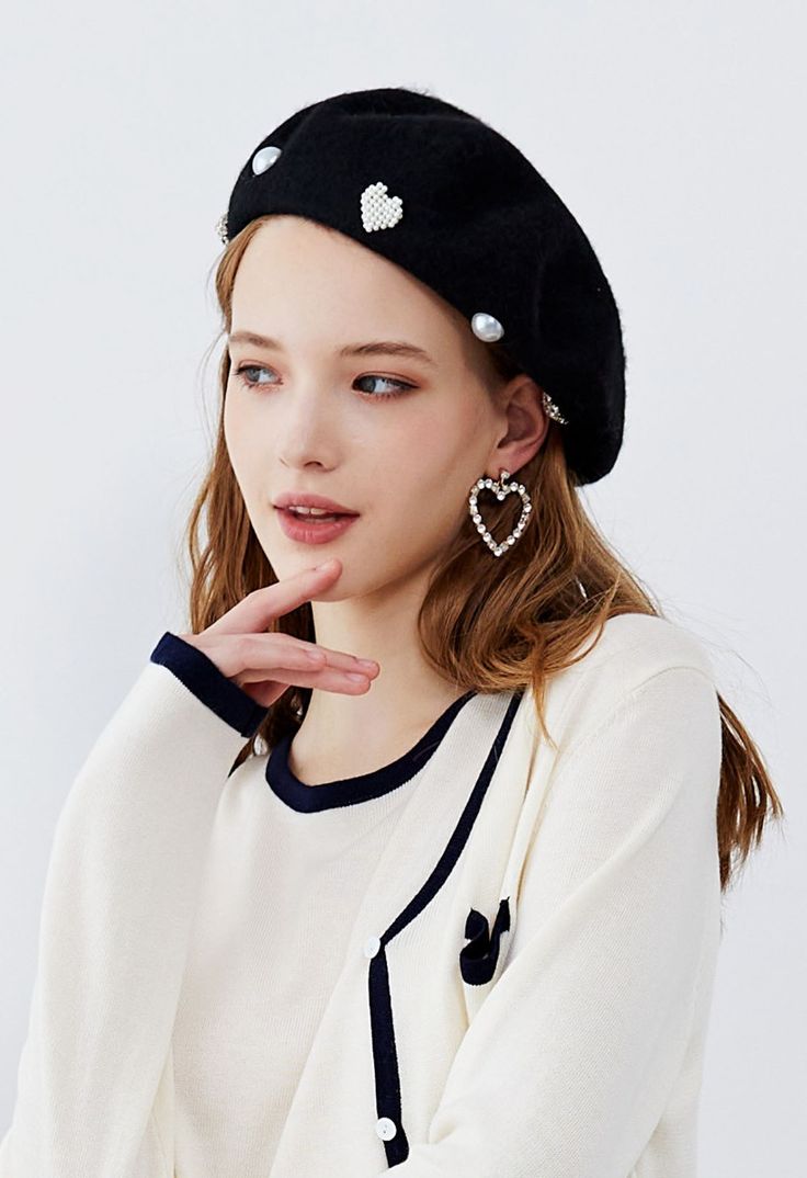 a woman wearing a black beret with white hearts on the front and back, standing in front of a white background