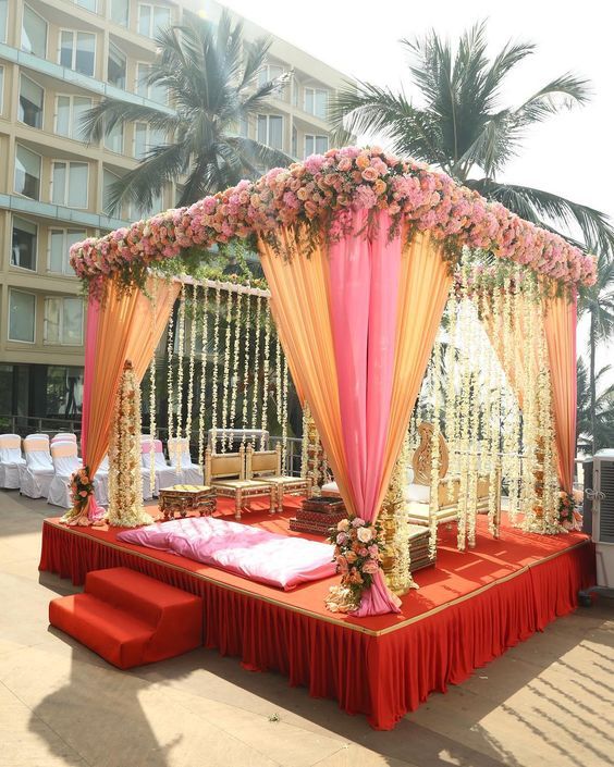 an outdoor wedding setup with red and orange drapes, flowers on the bed, and white chairs