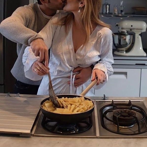 a man and woman are kissing in the kitchen while cooking food on the stove top