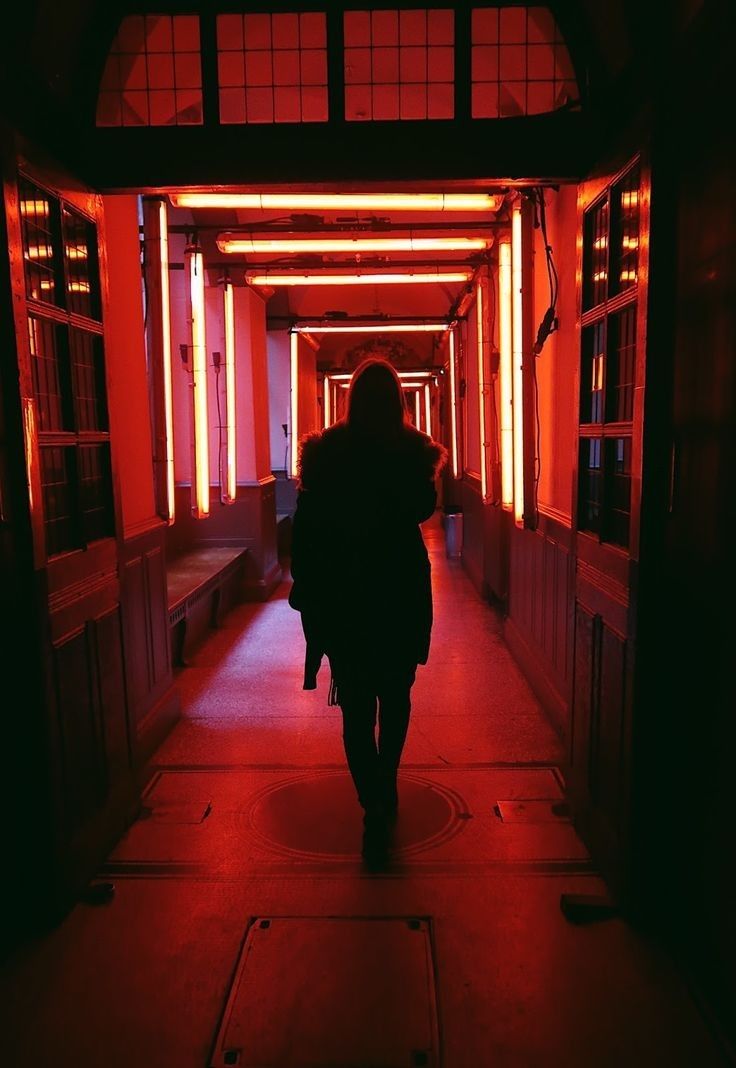 a person walking down a long hallway with red light coming from the ceiling and windows