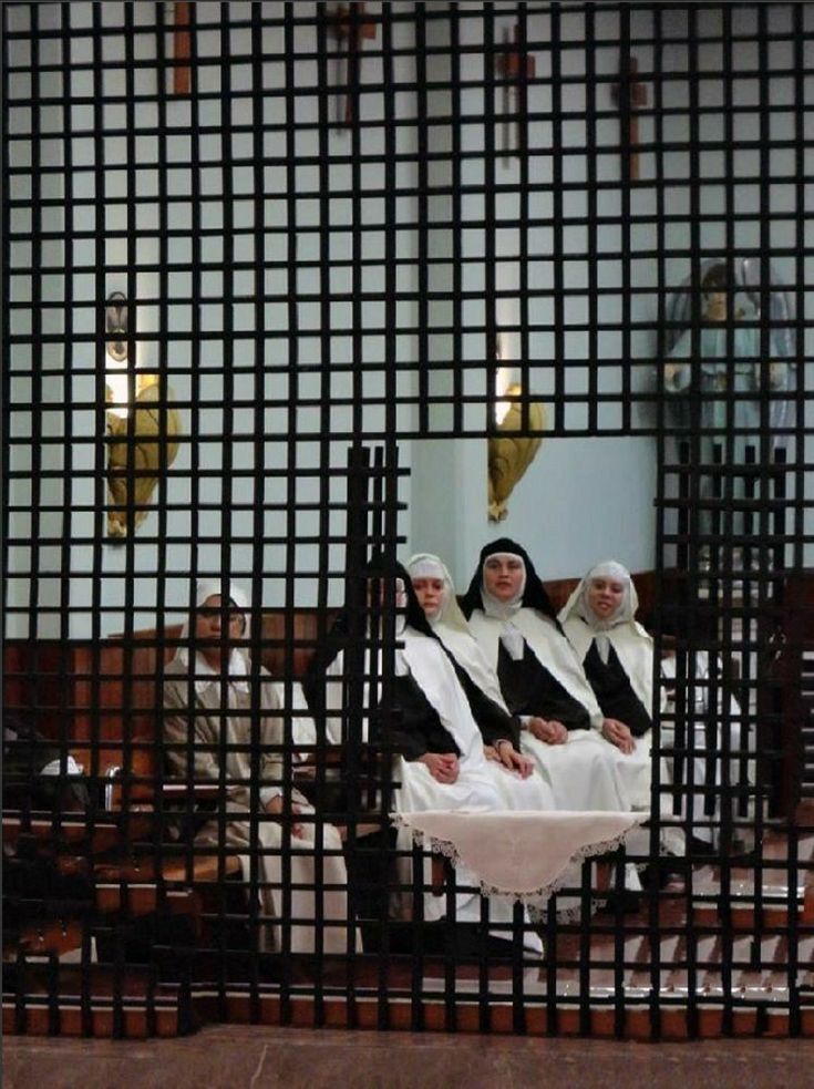 three women in white robes sitting on a bench behind a metal fence and looking at the camera