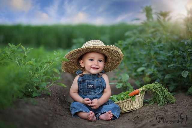 a baby wearing a straw hat sitting in the middle of a field with carrots