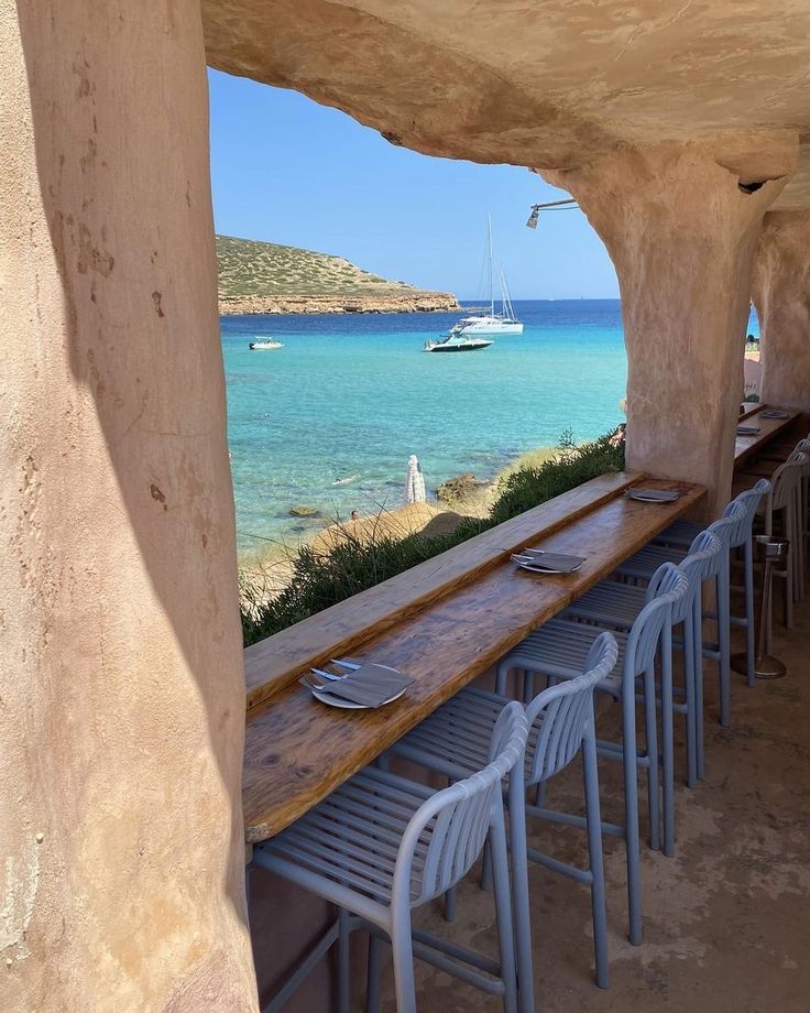 a long wooden table sitting under a stone arch next to the ocean with boats in the water