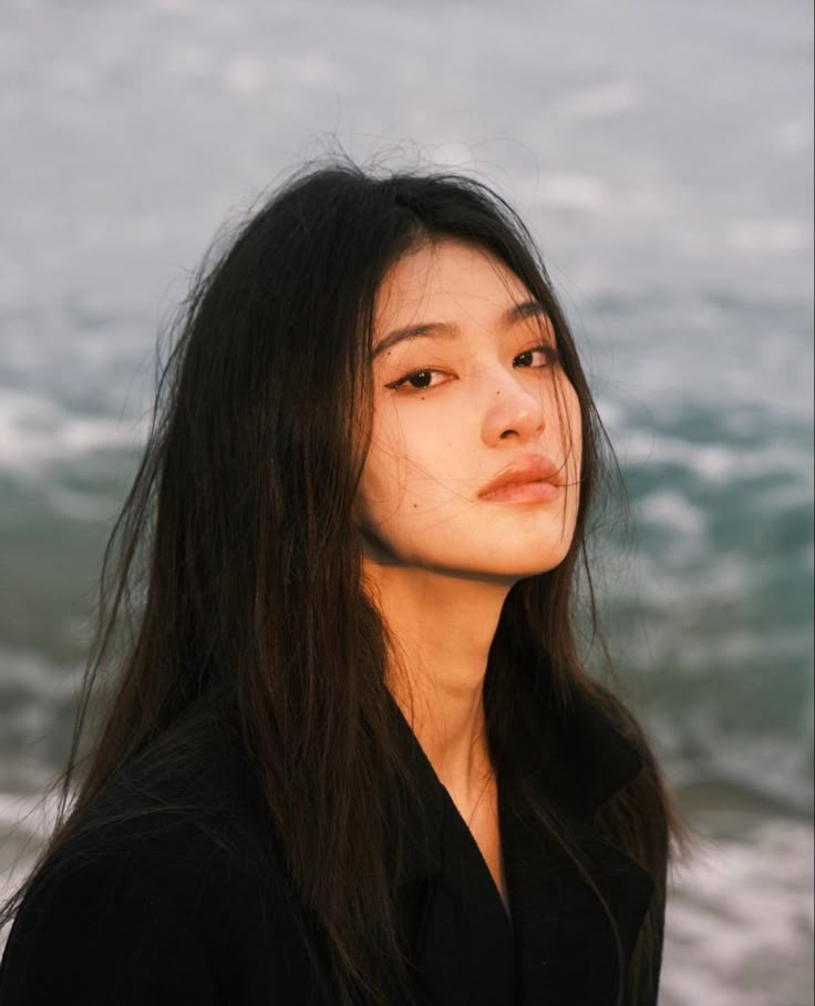 a woman with long black hair standing in front of the ocean and looking off into the distance