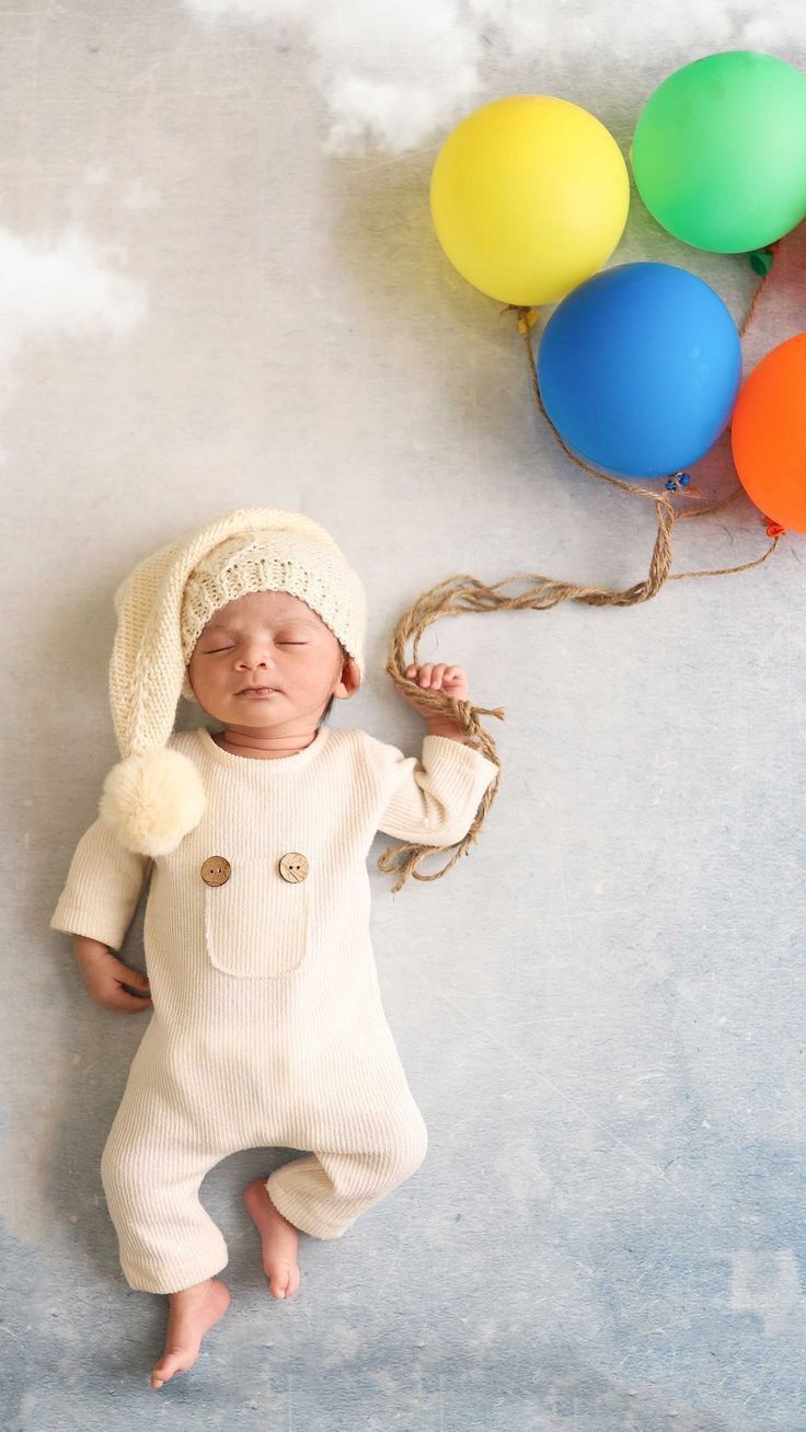 a baby is laying on the ground with balloons in the air behind him and wearing a knitted hat