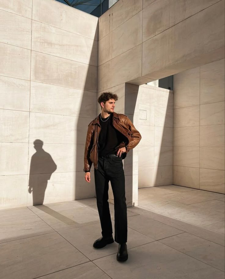 a man standing in front of a wall with his hands on his hips wearing a brown leather jacket and black pants