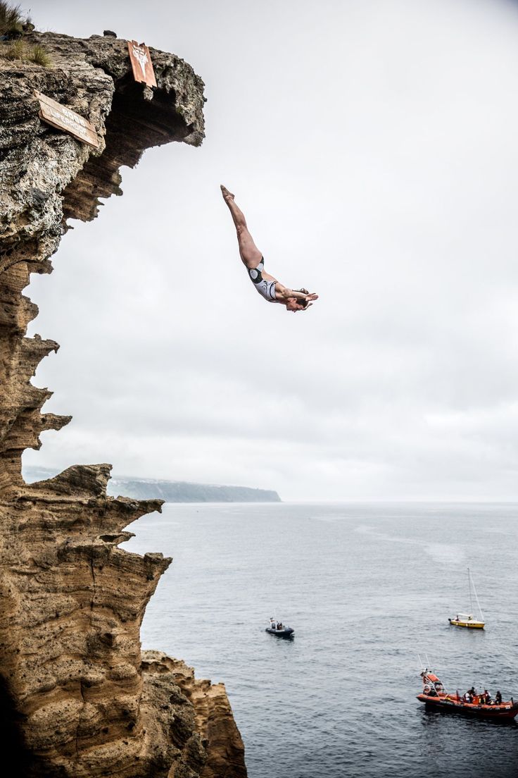 a man diving into the ocean from a cliff near a small boat in the water