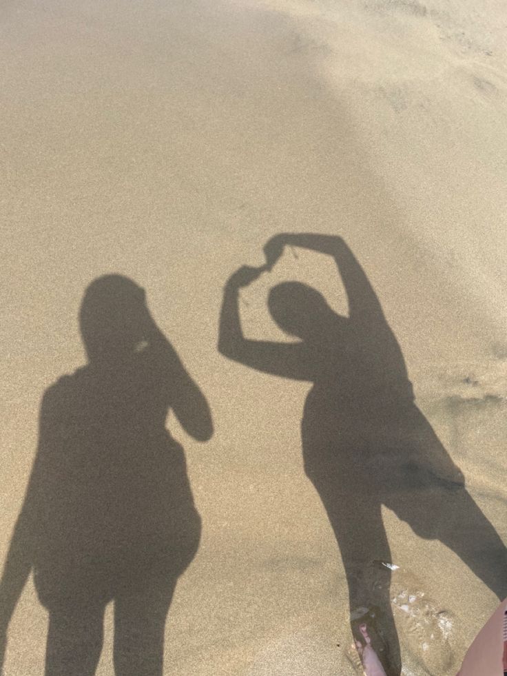 two people standing in the sand with their shadow on the beach and one person making a heart shape