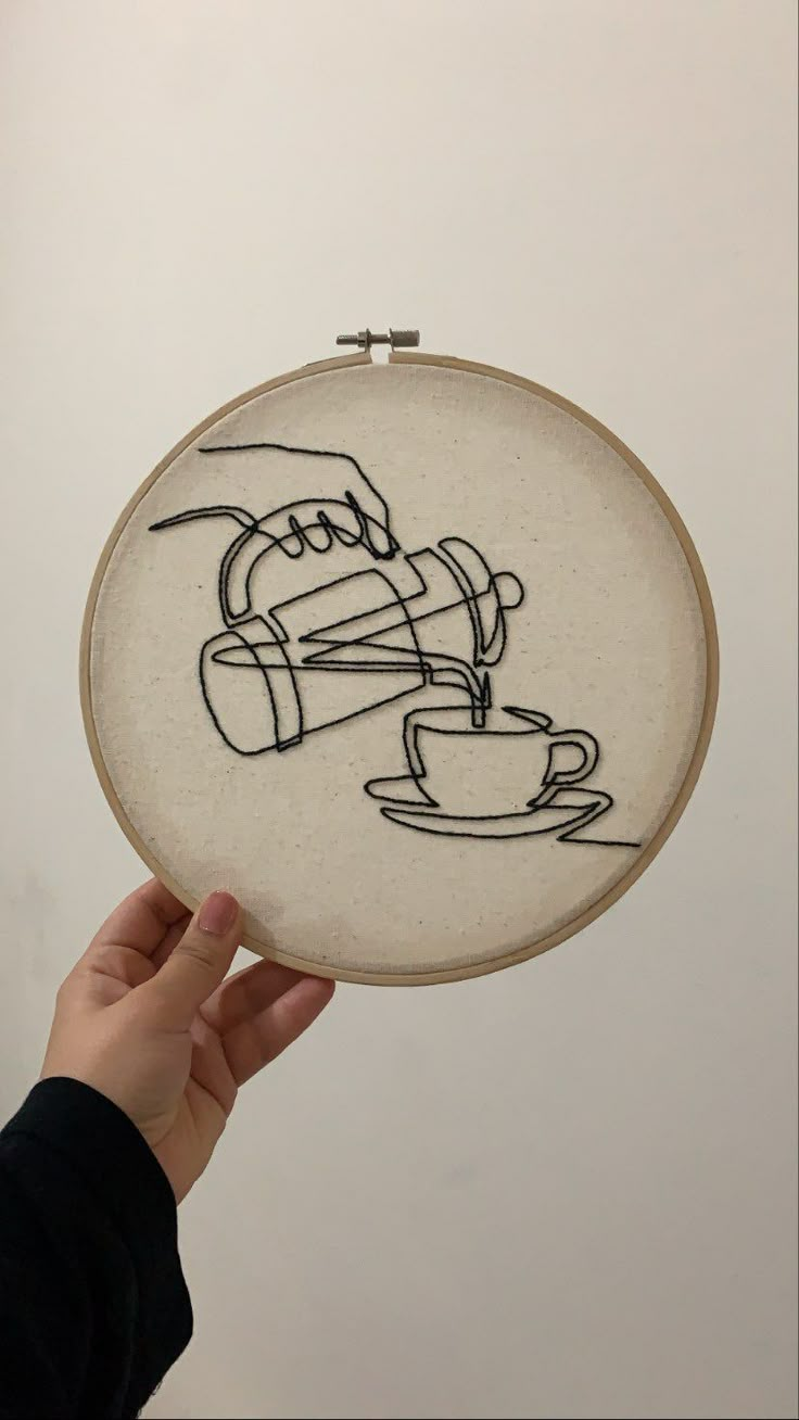 someone is holding up a embroidery art piece with a coffee cup and chair drawn on it