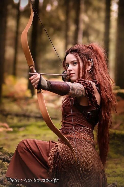 a woman with long red hair is holding a bow and arrow in the middle of a forest