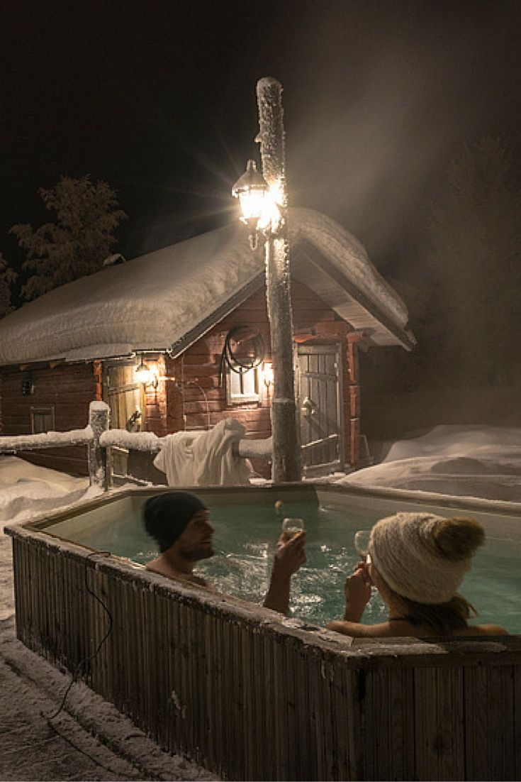 two people sitting in a hot tub with snow on the ground and a cabin behind them