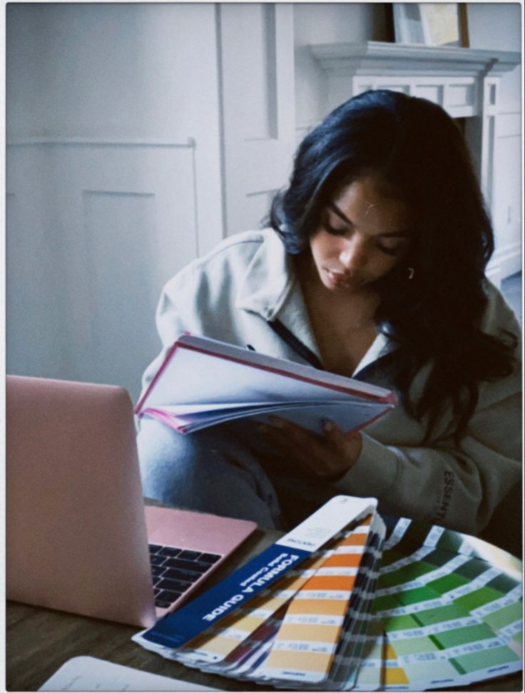 a woman sitting in front of a laptop computer holding a book and looking at it