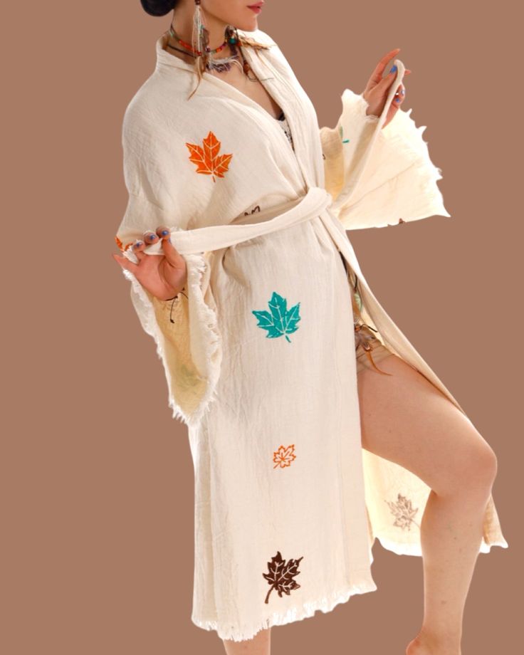 Wrap yourself in the warmth of the season with the Autumn Breeze Kimono. This lightweight, cream-colored kimono is beautifully adorned with vibrant maple leaves in earthy hues. Its soft, natural fabric and relaxed silhouette make it the perfect layering piece for transitional weather. Whether you’re enjoying a cool evening outdoors or adding an artisanal touch to your everyday look, this kimono brings an effortless sense of style. With wide sleeves and a matching waist tie, this one-size-fits-al White Cotton Kimono For Fall, Long White Kimono For Fall, White Long Kimono For Fall, Fall Beige Cotton Kimono, Cream Long Sleeve Fall Kimono, Beige Kimono For Beach In Fall, Beige Bohemian Kimono For Fall, Beige Bohemian Kimono For Loungewear, Bohemian Beige Kimono For Fall