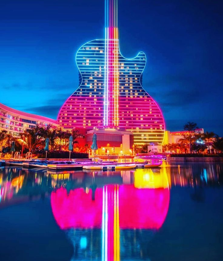 the guitar shaped building is lit up in pink and blue lights, reflecting on the water