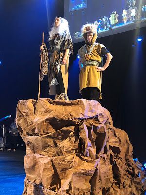 two people standing on top of a rock in the middle of a stage with lights behind them