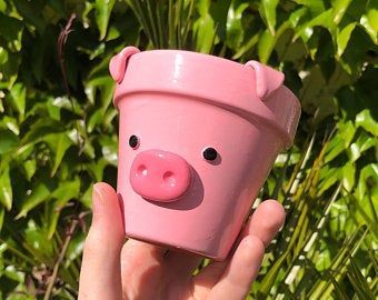 a hand holding a pink cup with a pig face on it's side and green plants in the background