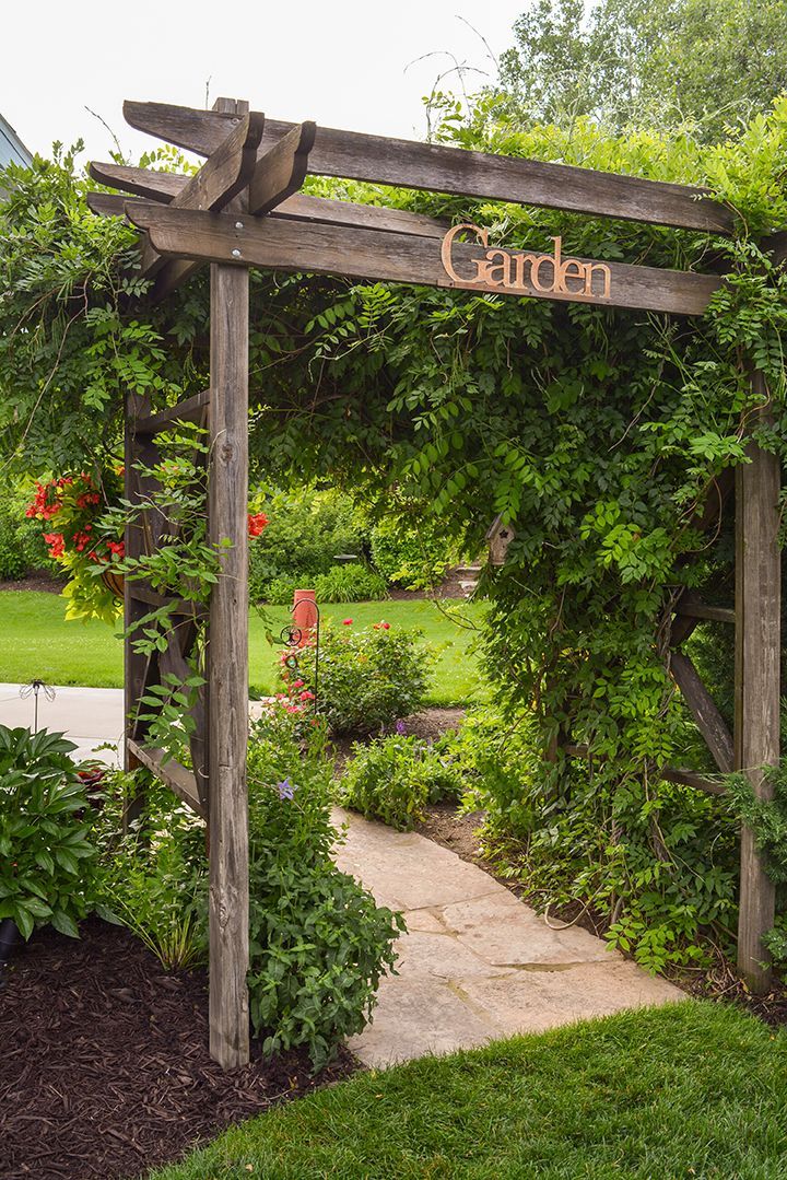 a wooden garden arbor with the word garden on it's side in front of some bushes and flowers