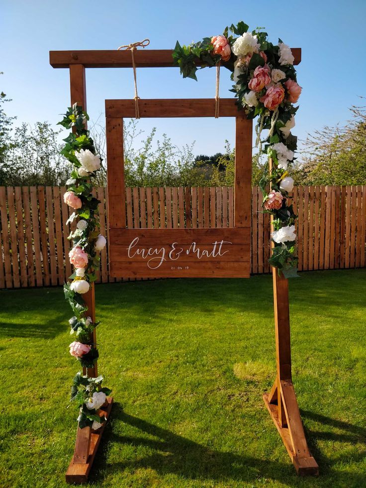 an outdoor wedding ceremony setup with flowers and greenery on the grass, in front of a wooden frame