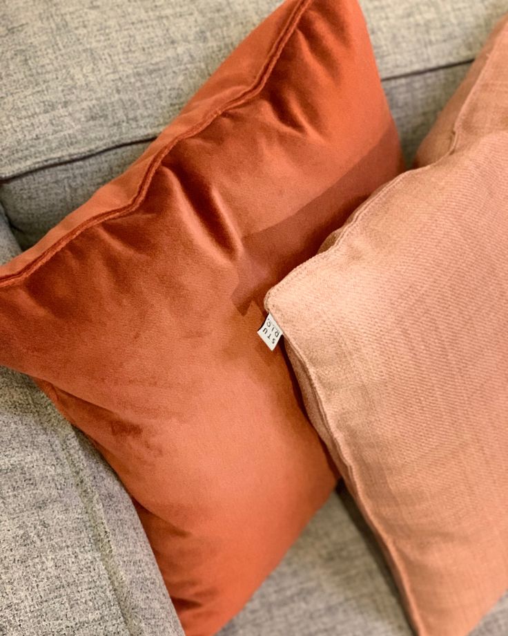 two orange pillows sitting on top of a gray couch next to a pillow with a white tag