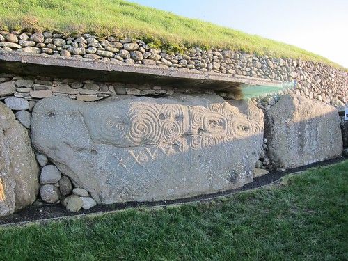 a stone wall with carvings on it and grass growing on the top, in front of a grassy hill