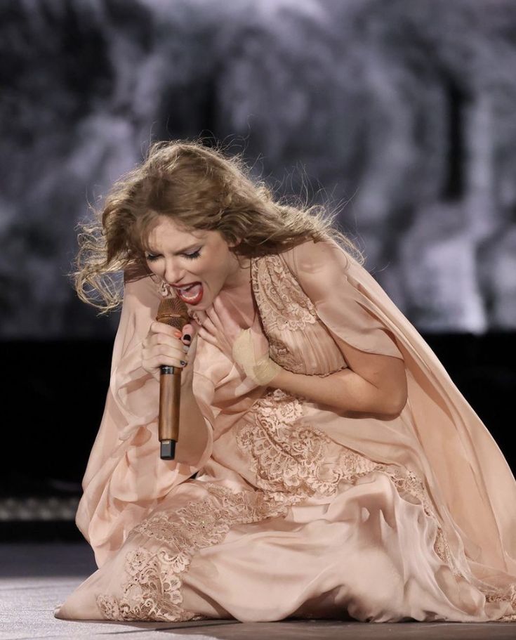 a woman kneeling down holding a microphone in her right hand and wearing a pink dress