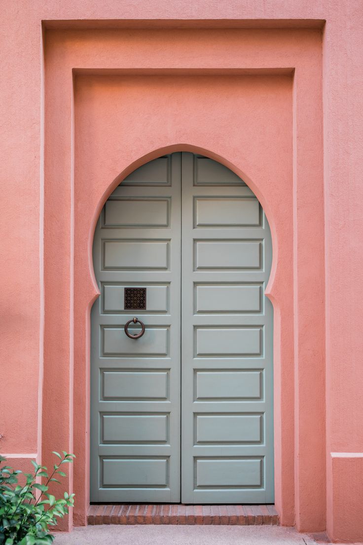 an arched doorway with a door handle on the side of a pink building that has green shutters