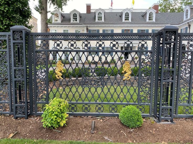 an iron gate in front of a large house with trees and bushes around it on the lawn