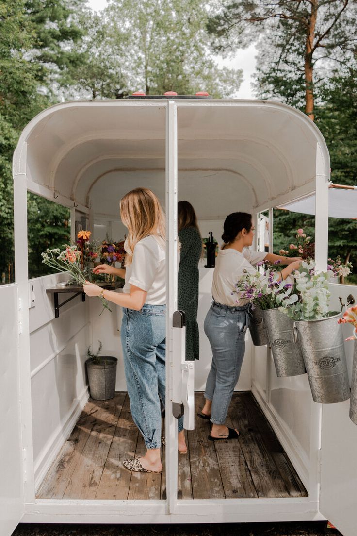 two women are standing in the back of a white truck with flowers on it's side