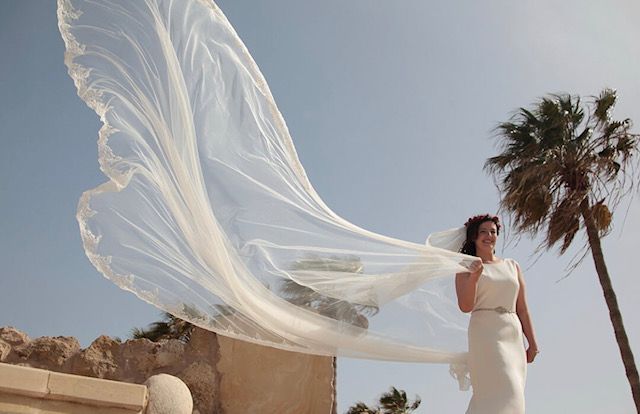 a woman in a white wedding dress holding a veil over her head while standing next to a palm tree