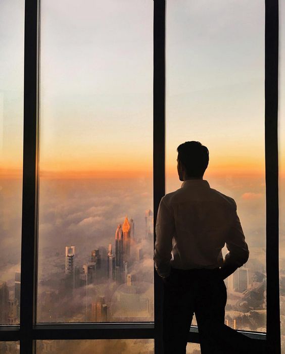 a man standing in front of a window looking out on the city skyline at sunset