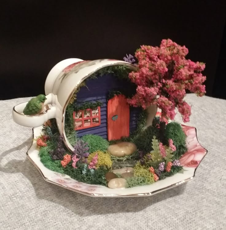 a tea cup with a house in the middle and flowers around it on a table