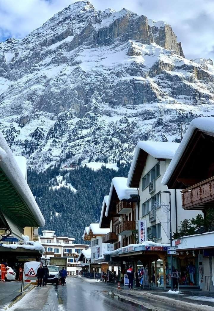 people are walking down the street in front of snow covered mountains and buildings with shops on either side