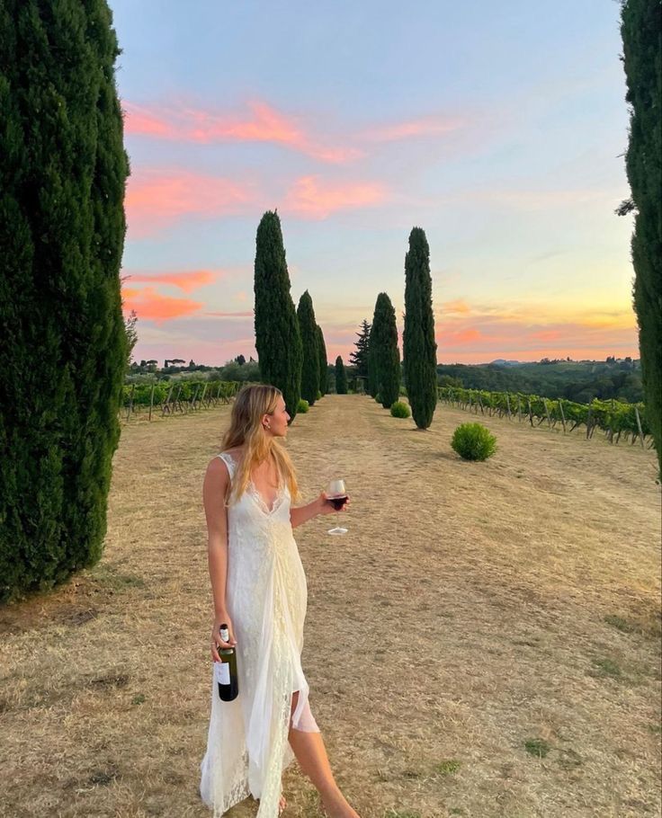 a woman in a white dress holding a glass of wine and standing on a dirt road