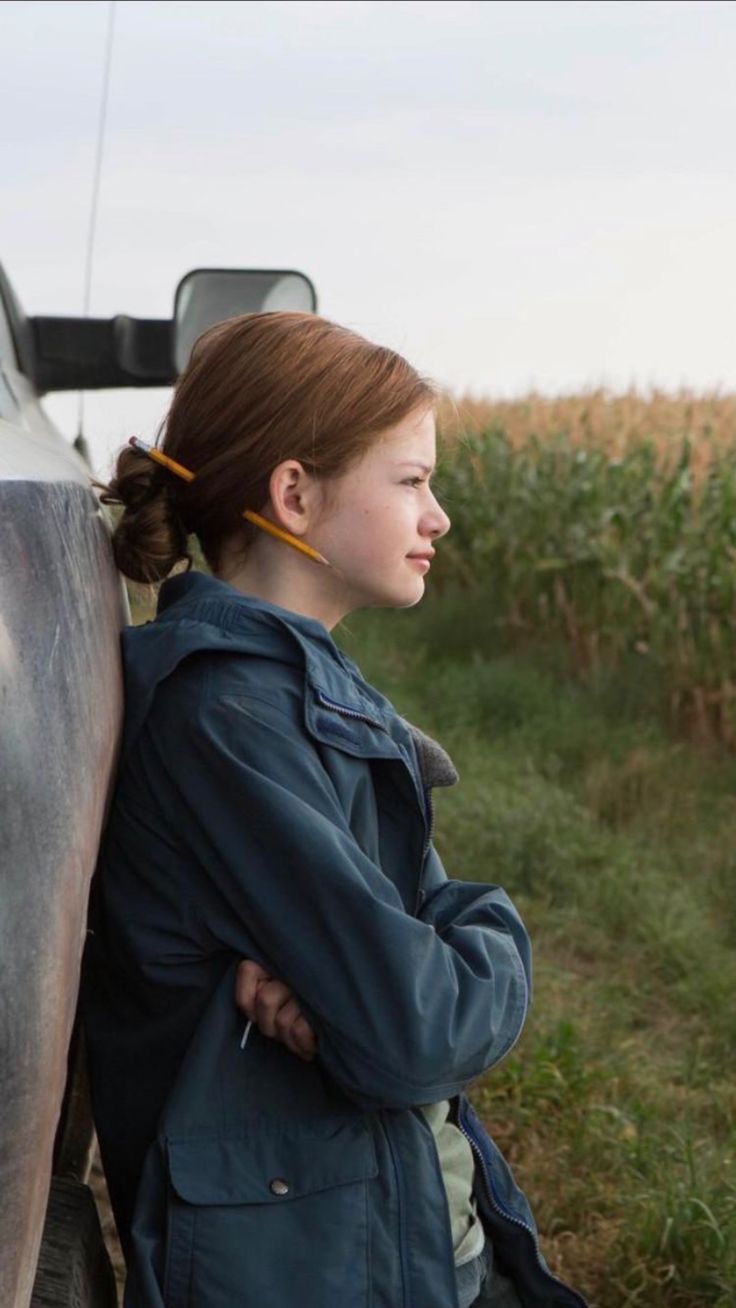 a woman leaning against the side of a truck in a corn field with her back to the camera