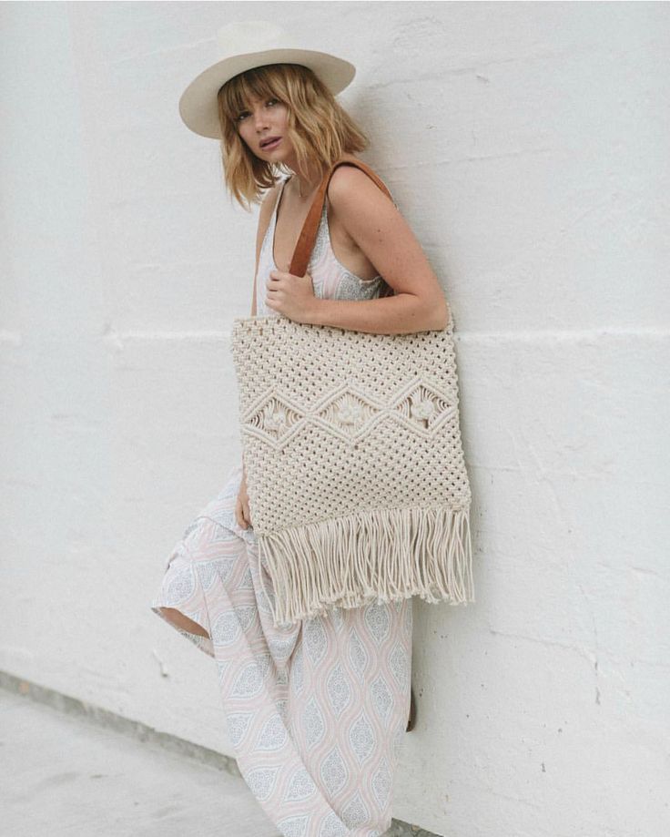 a woman leaning against a white wall holding a beige bag with fringes on it