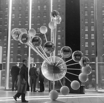 people standing in front of a building with large balls on the floor and one man walking past it