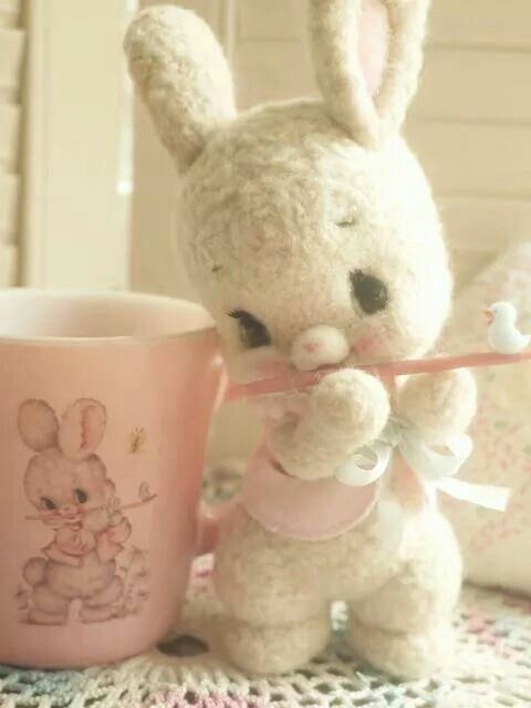 a stuffed rabbit holding a pink cup on top of a table next to a mug