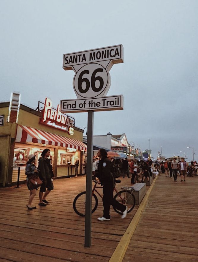 people walking and riding bikes on a boardwalk next to the street sign that says santa monica 66 end of the trail